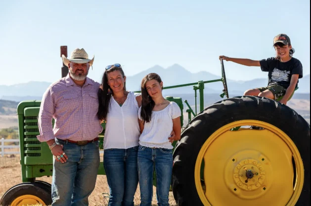 The Bennett family standing with their son on a green John Deere tractor, Colorado mountains in the background